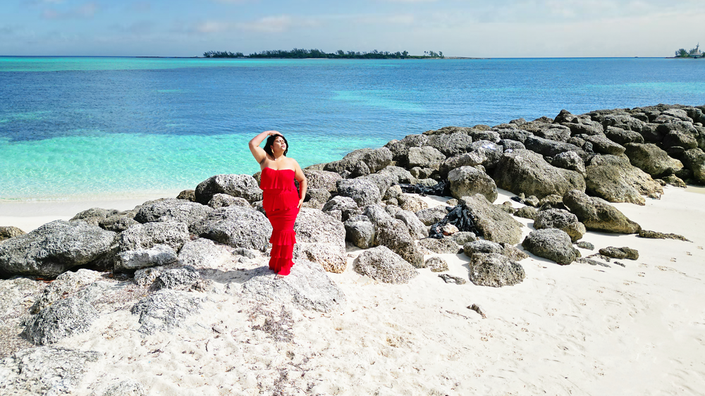 A person in red stands on rocky coastal outcrops overlooking crystal clear turquoise waters.