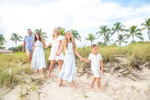 Family of five posing on a sandy beach path surrounded by palm trees and green dunes in matching white outfits.