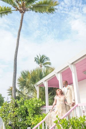 Couple in wedding attire descends pink stairs of tropical venue with palm trees and blue sky backdrop.