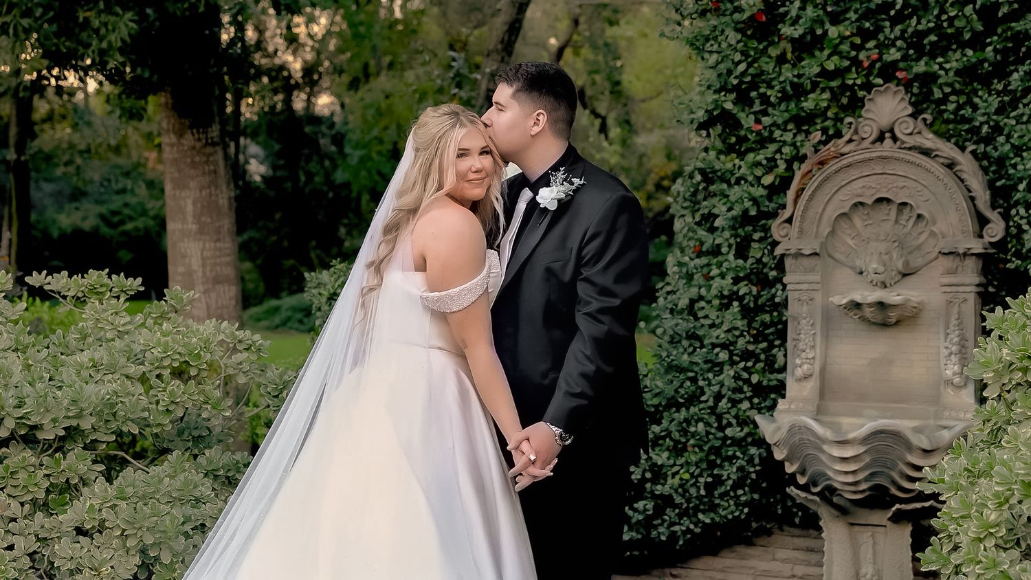 Bride and groom share a romantic kiss in a lush garden near a stone fountain, bride in off-shoulder ballgown.