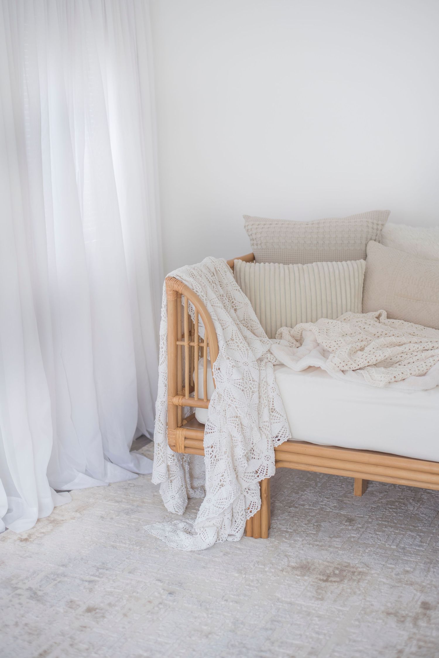 Cozy daybed with natural wood frame adorned with cream pillows and lace throw blanket near white flowing curtains.