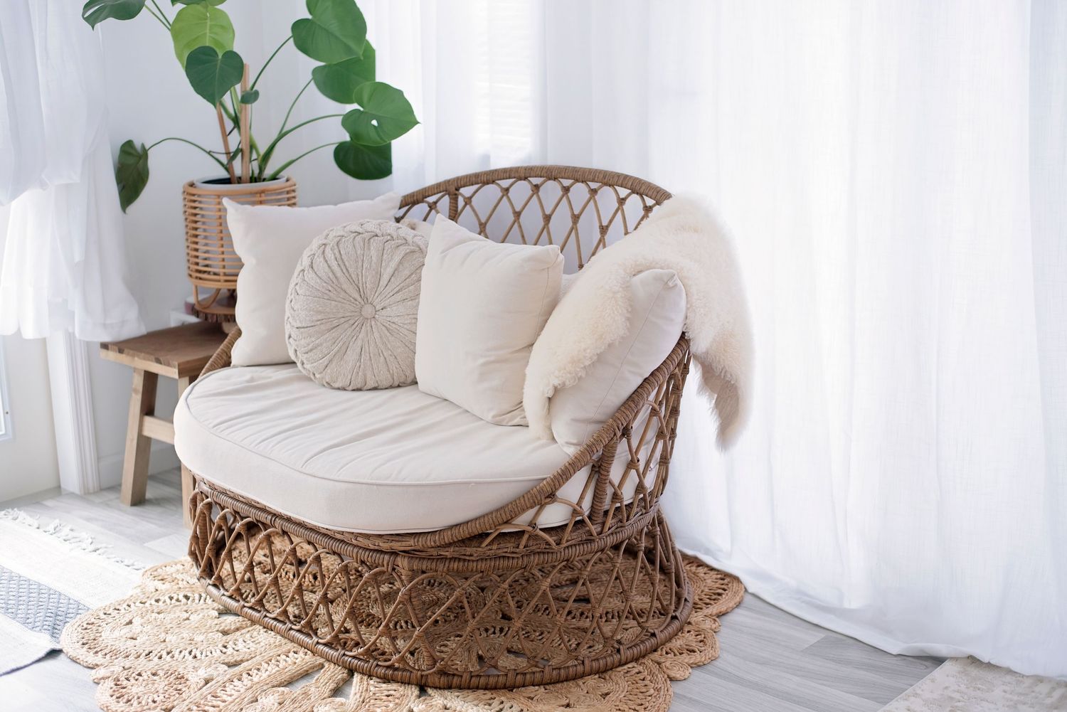Cozy rattan papasan chair with plush white cushions and decorative pillows sits beside a houseplant in a bright room.
