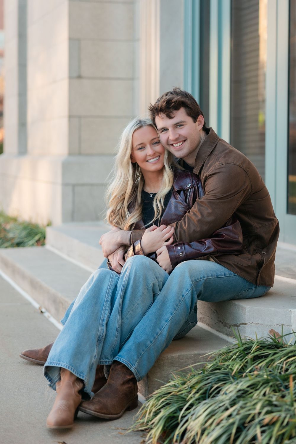 A couple sits close together on concrete steps outside a building, wearing casual denim and leather jackets.