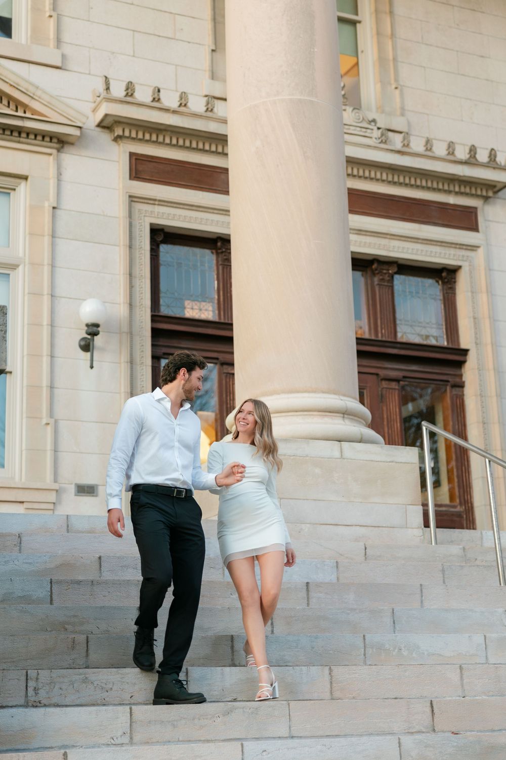 A couple in elegant white outfits walks down stone steps by a classical building with tall columns.