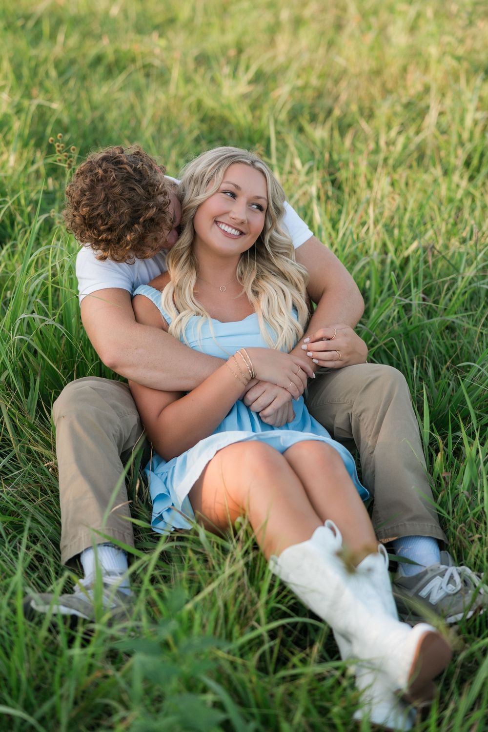 Two people cuddling together while sitting in tall green grass on a sunny afternoon.