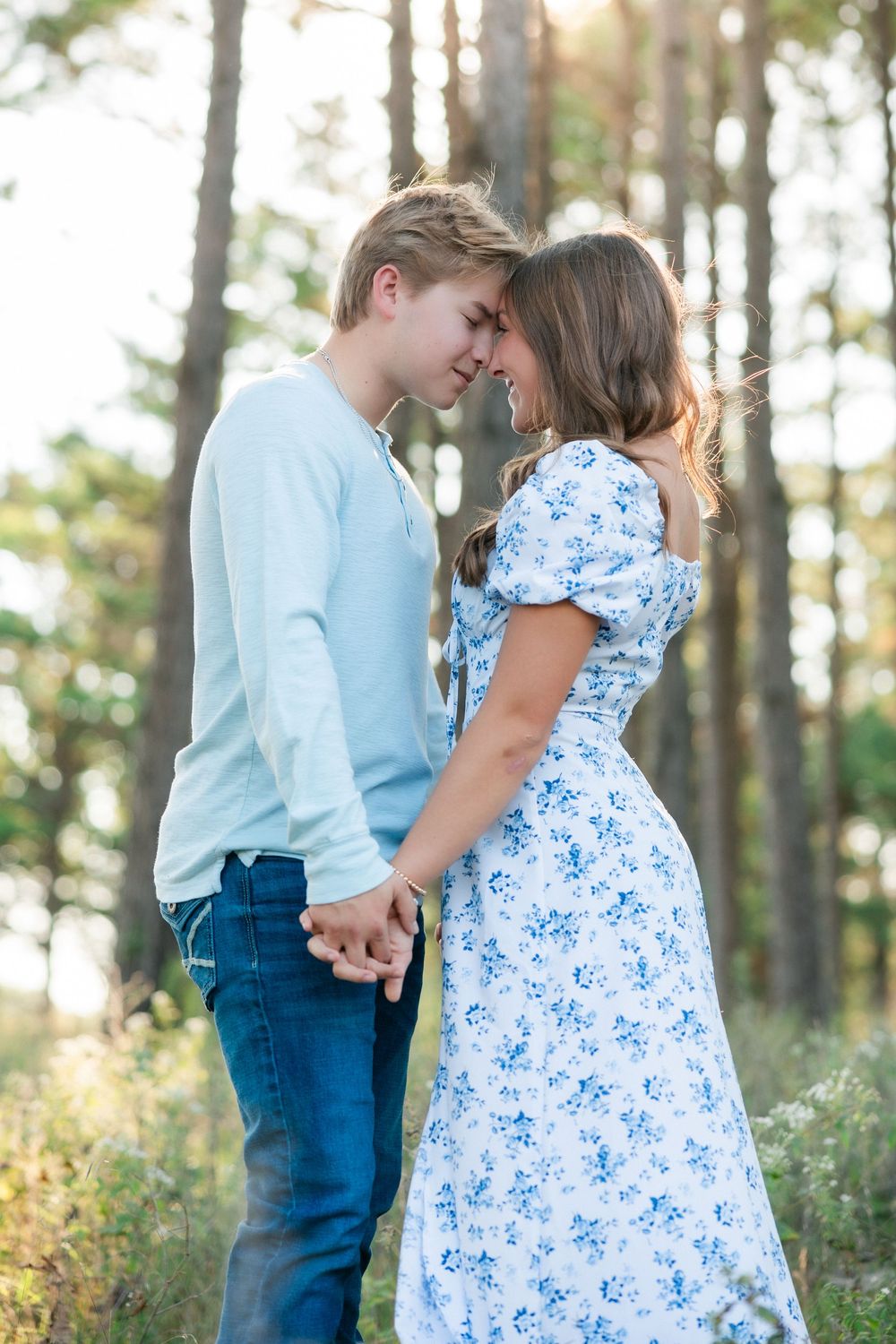 Romantic young couple in blue outfits sharing an intimate moment while holding hands in a sunlit forest setting.