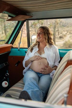 A pregnant woman sits comfortably in the passenger seat of a vintage turquoise car, enjoying a relaxed moment