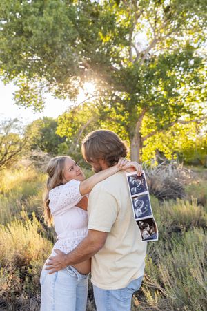 A couple embraces outdoors in Jacksonville North Carolina while holding ultrasound photos in a sunlit park with lush green trees