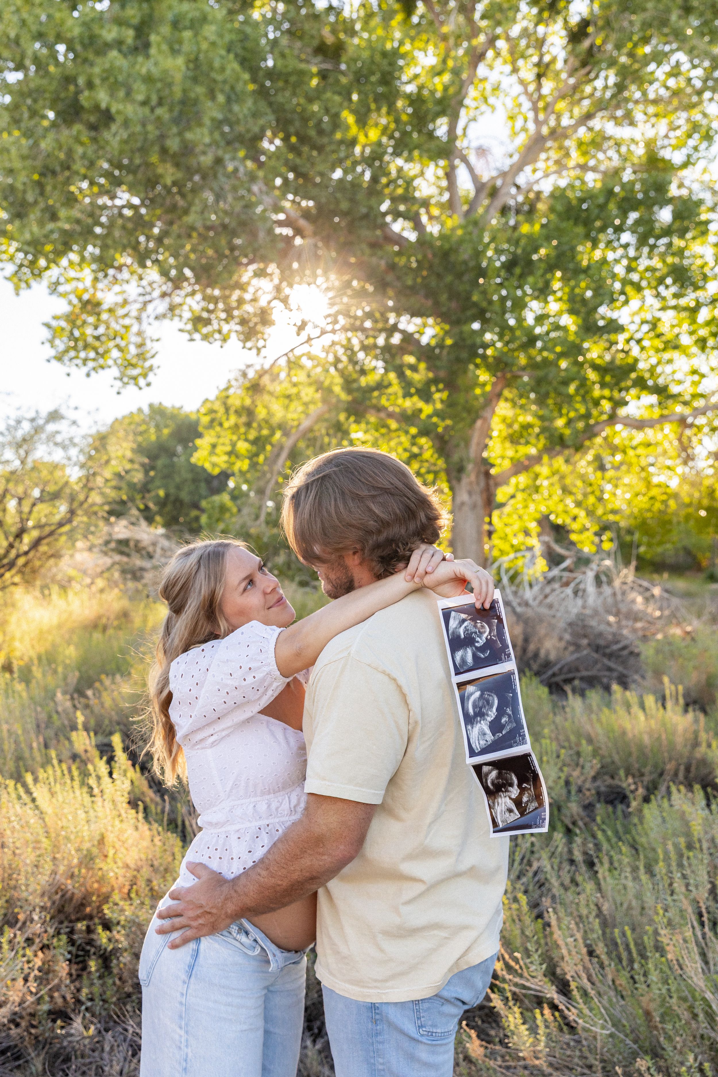 A couple embraces outdoors in Jacksonville North Carolina while holding ultrasound photos in a sunlit park with lush green trees
