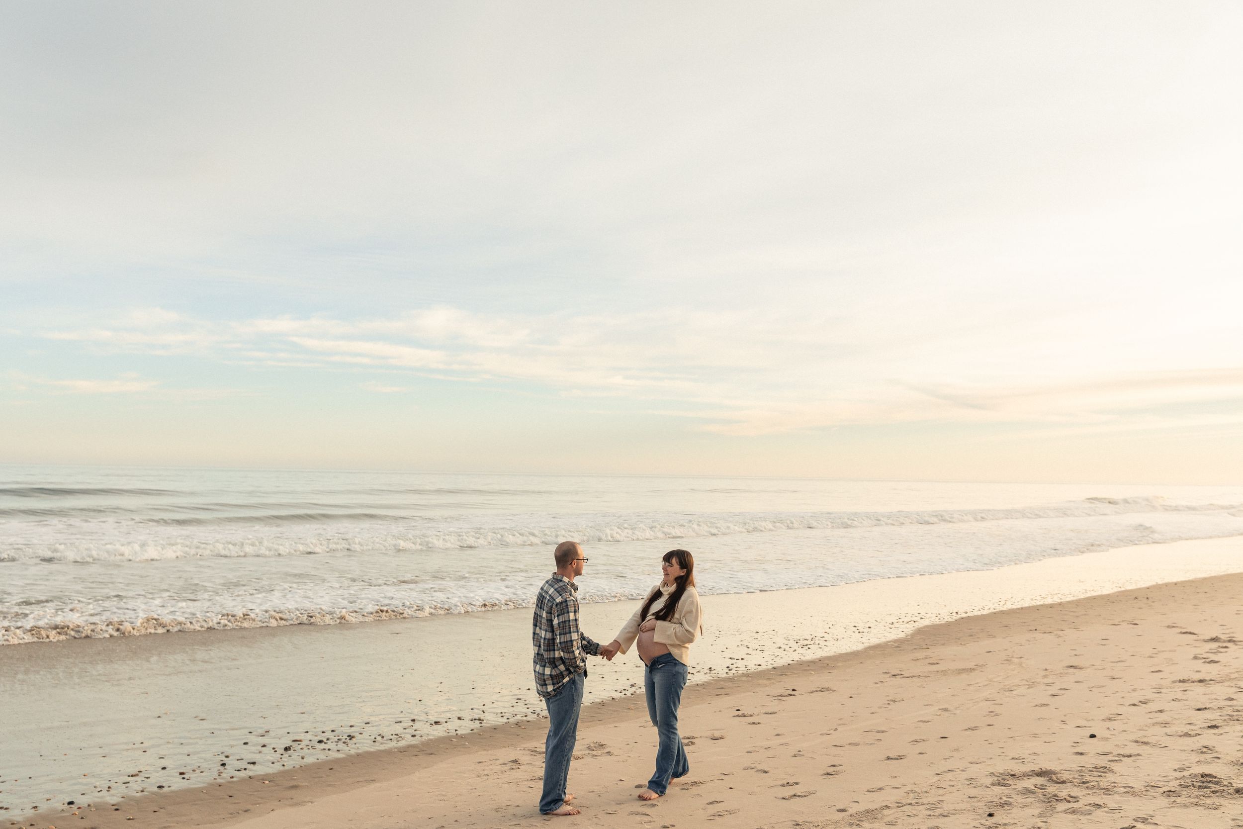 Coastal North Carolina Maternity Photographer, Couple holding hands on a sandy beach at sunset with ocean waves in the background.