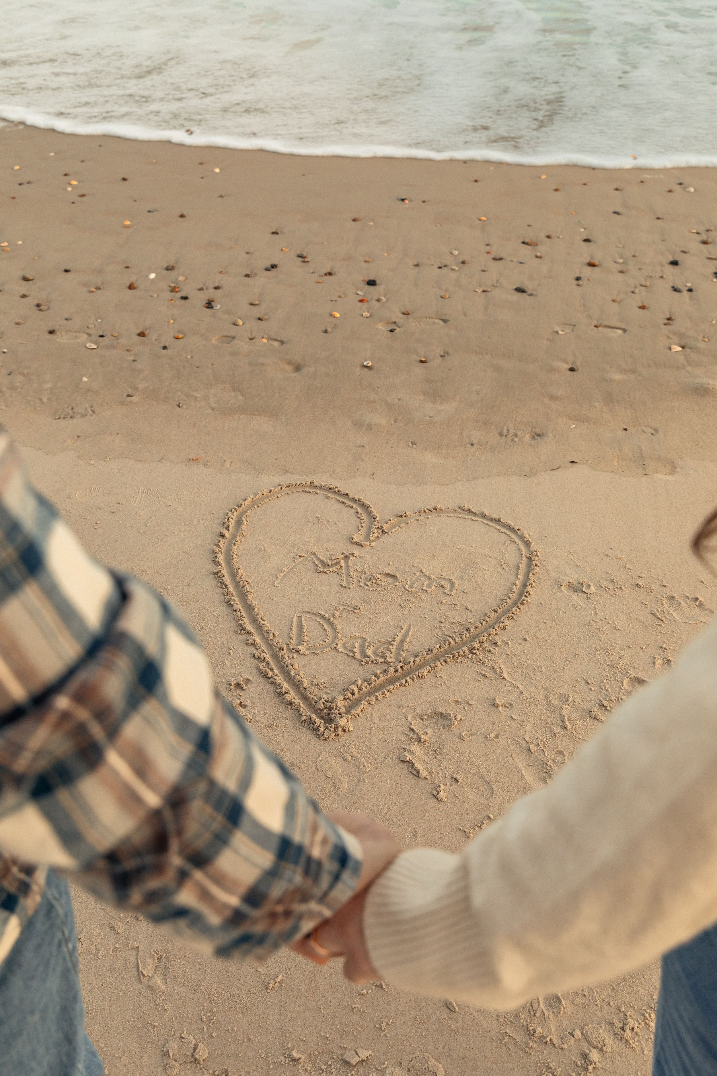 Couple holding hands on a beach in North Carolina with a heart drawn in the sand containing "mom + dad", with ocean waves in the background.