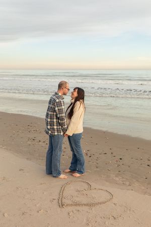 Couple holding hands on a sandy beach with a heart drawn in the sand, ocean waves in the background at sunset on North Topsail Beach in NC