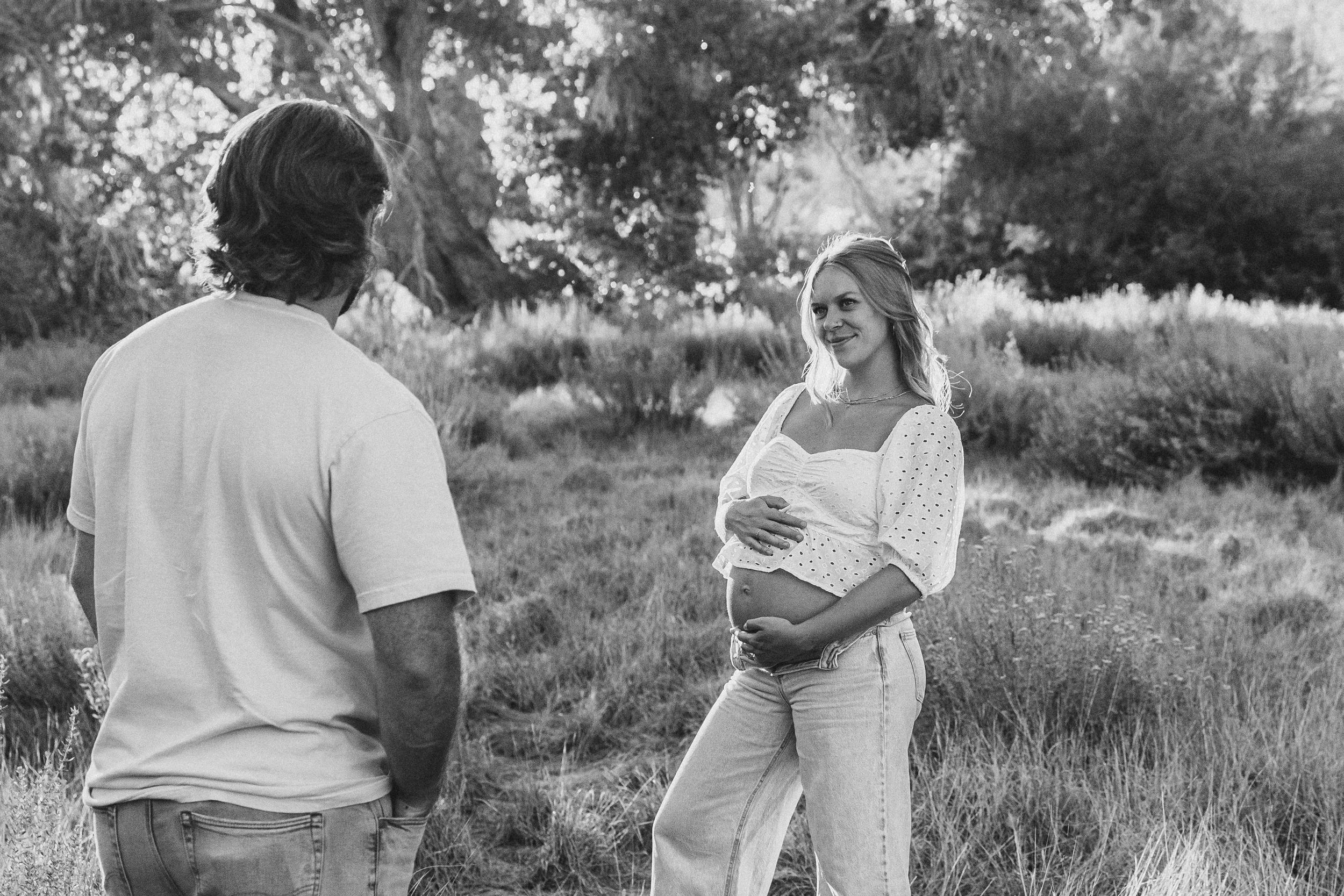 A soon to be mom standing in a grassy field in North Carolina while holding her belly and looking at her husband