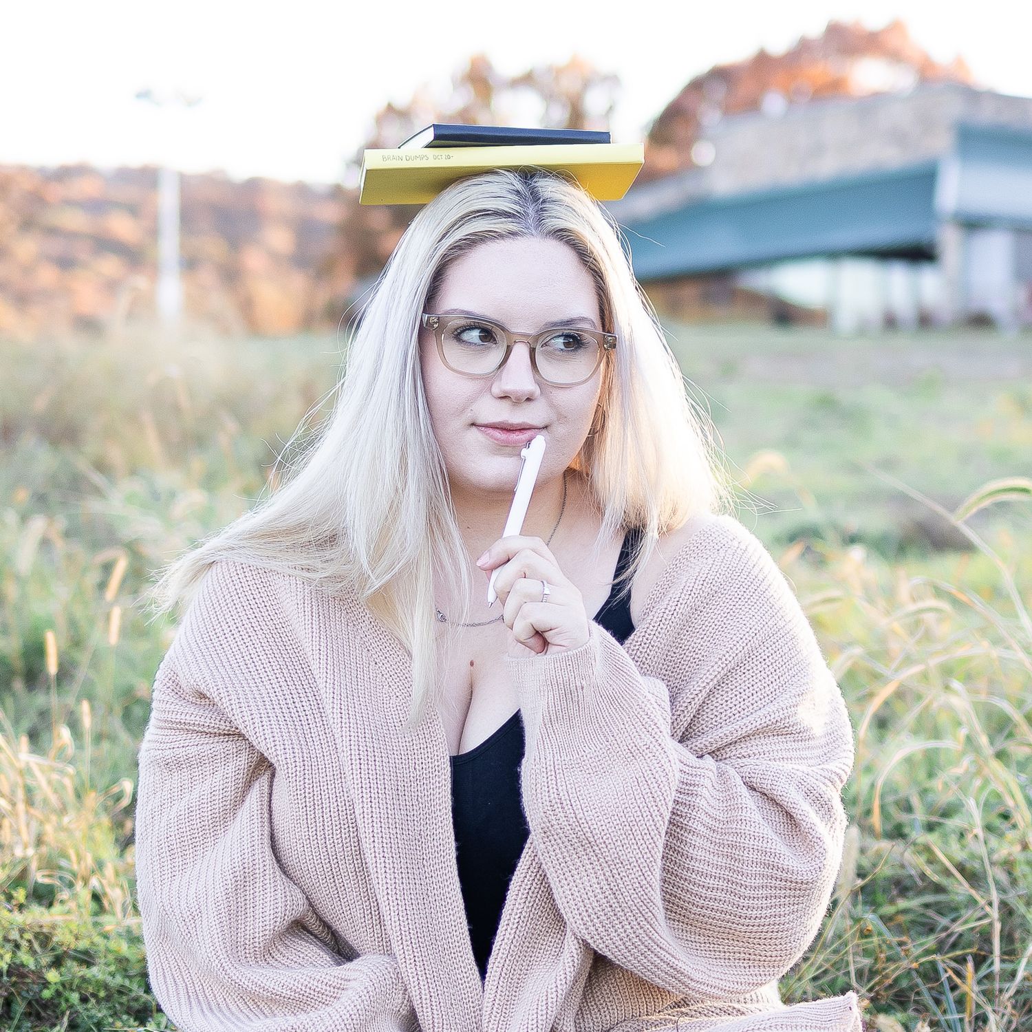 Someone wearing glasses and chunky beige sweater poses with finger to lips in outdoor setting.