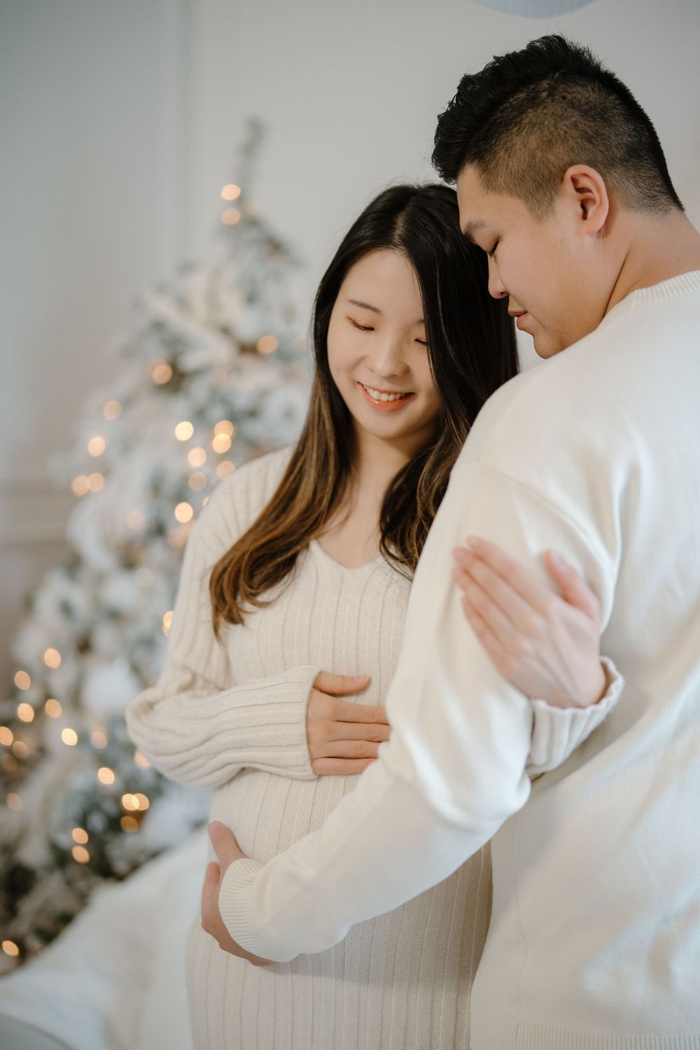 A romantic couple in white sweaters embraces in front of a glowing Christmas tree in a dreamy winter setting.