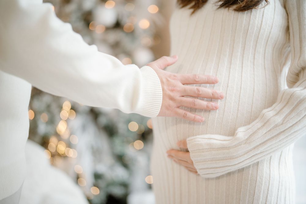 Elegant bride in a flowing white dress cradles her baby bump in a dreamy winter scene with soft twinkling lights.
