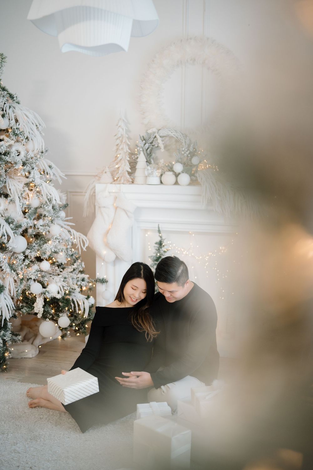 A couple in black clothing sits together near a decorated Christmas tree and white fireplace mantel with festive decor.
