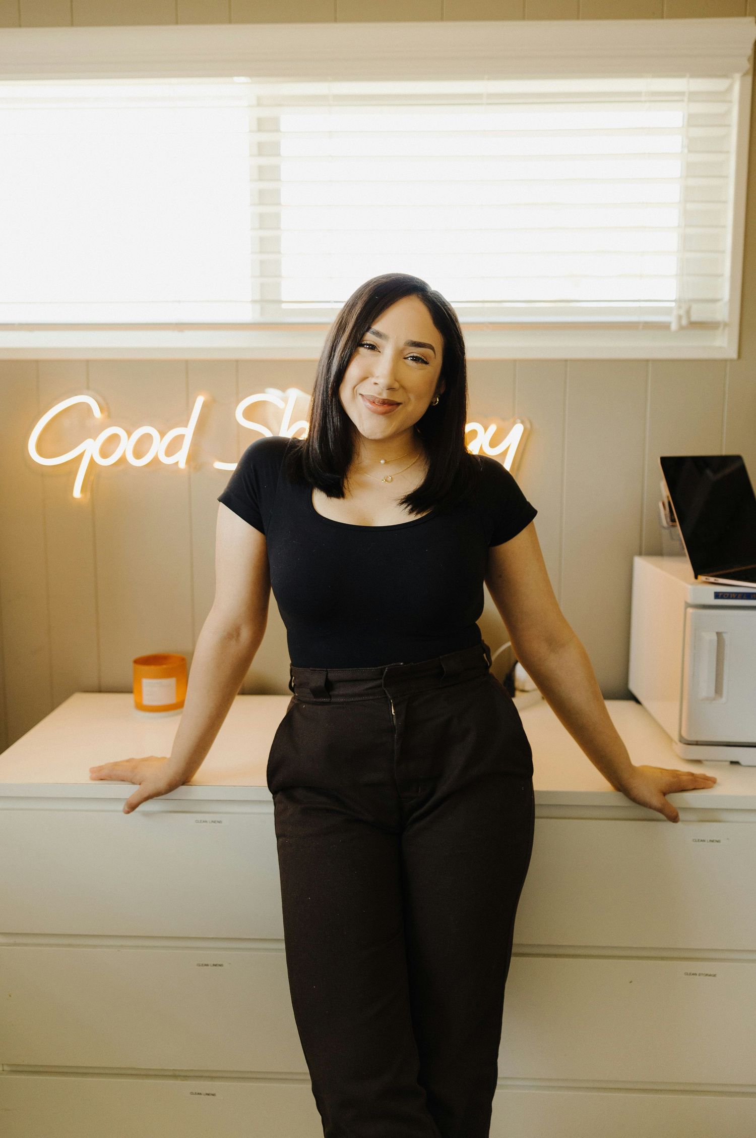 Person poses in front of neon sign wearing black outfit in minimalist office setting.
