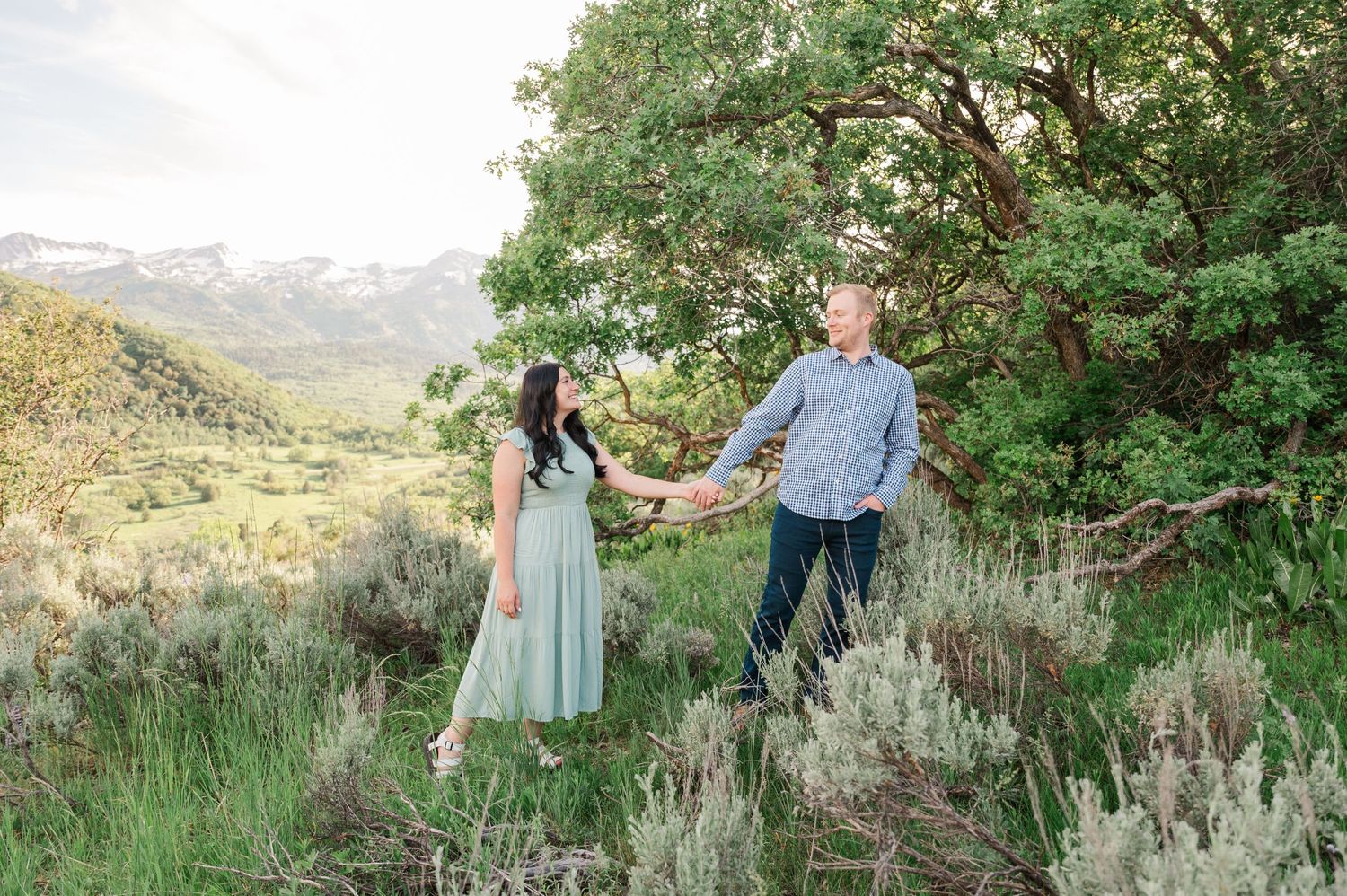 A couple poses together in a natural setting with mountains and sagebrush in the background.