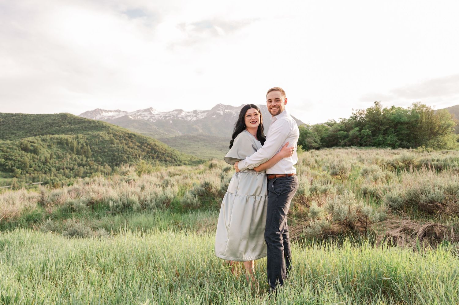 A couple embraces while standing in a meadow with mountain views during sunset.