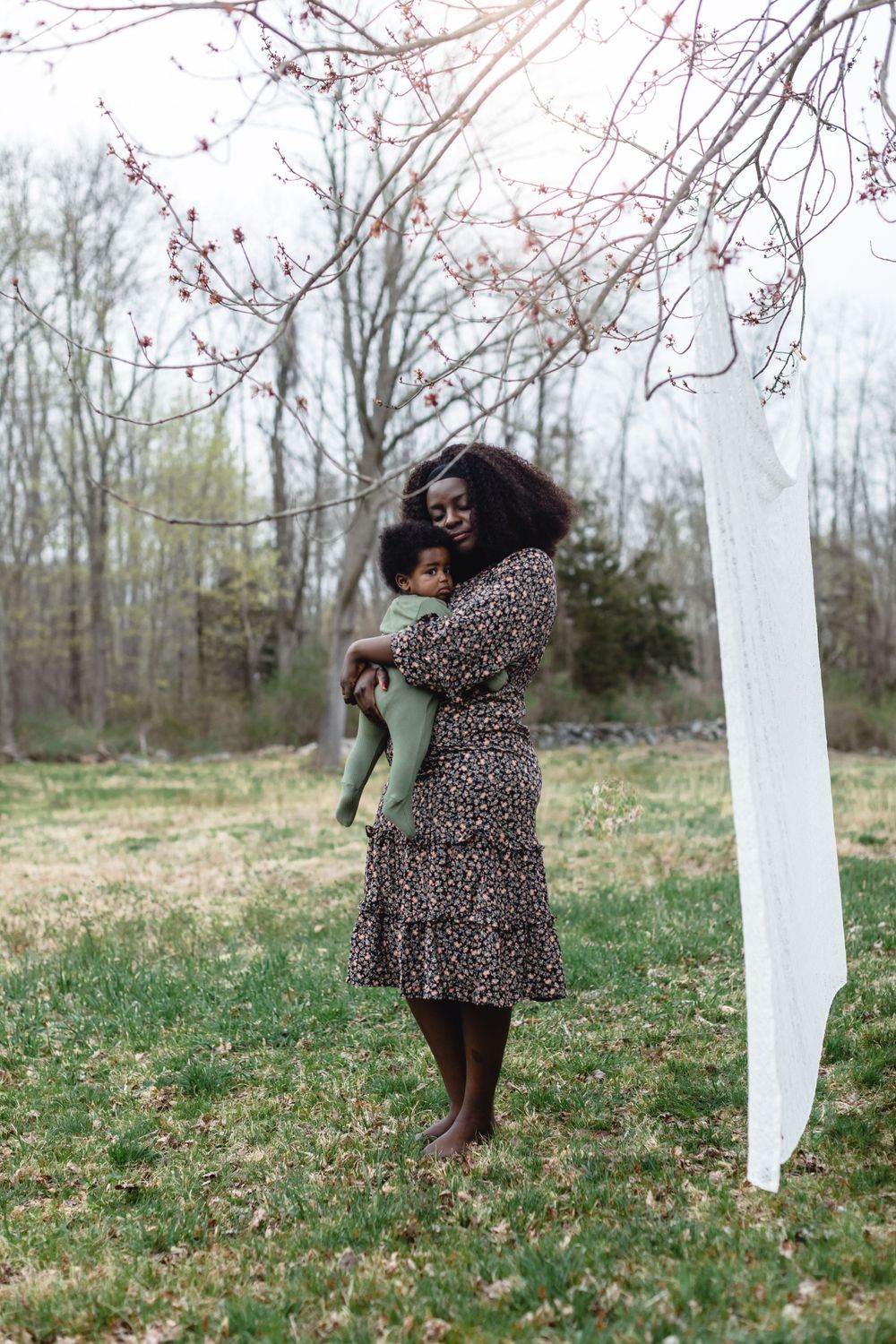 A mother wearing a floral dress stands in a field under flowering trees while holding her baby boy in Concord, MA for a motherhood session.