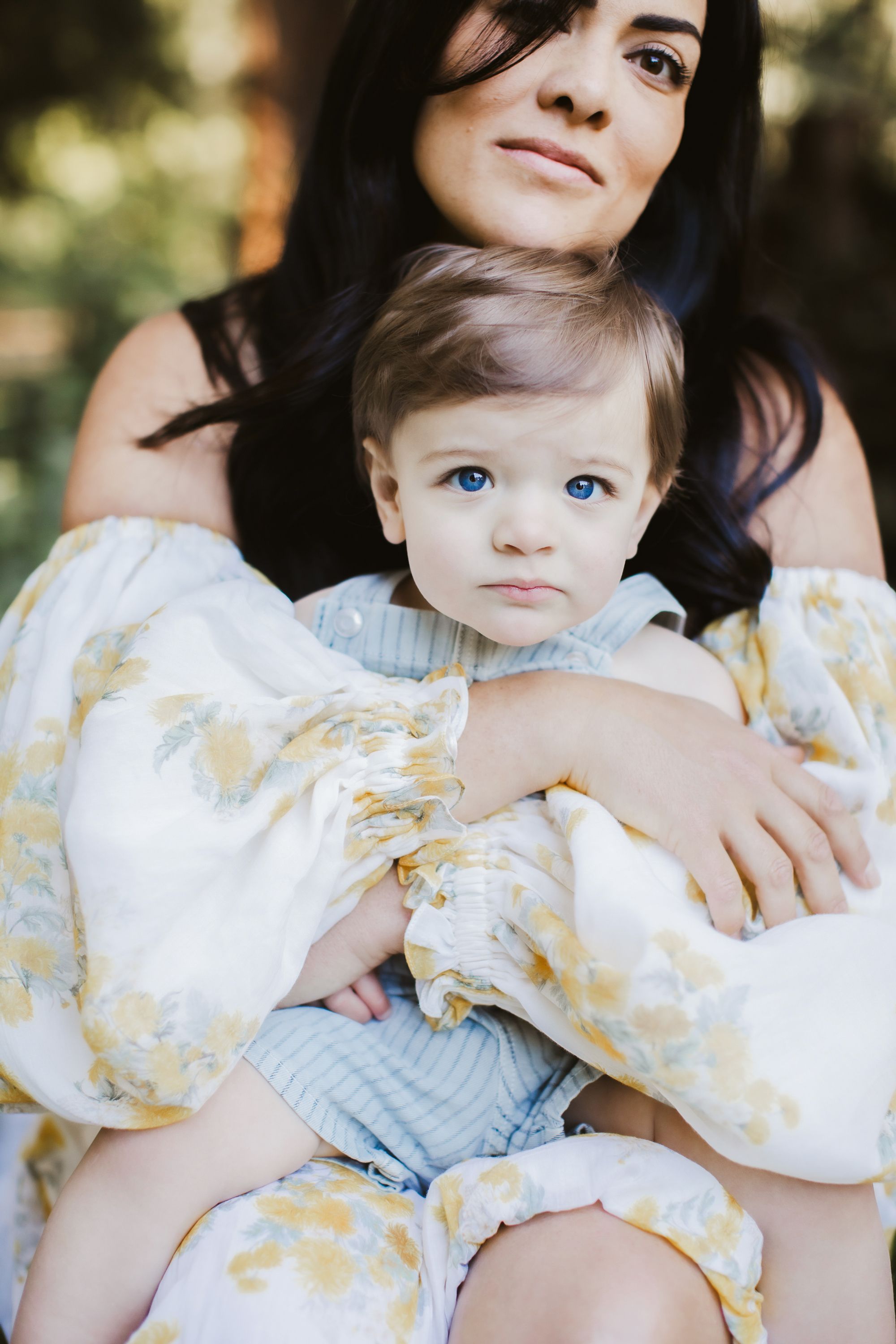 Mother with dark hair wearing a puff sleeve white flora; dress embraces her one year old, blue eyed boy for motherhood family photos.