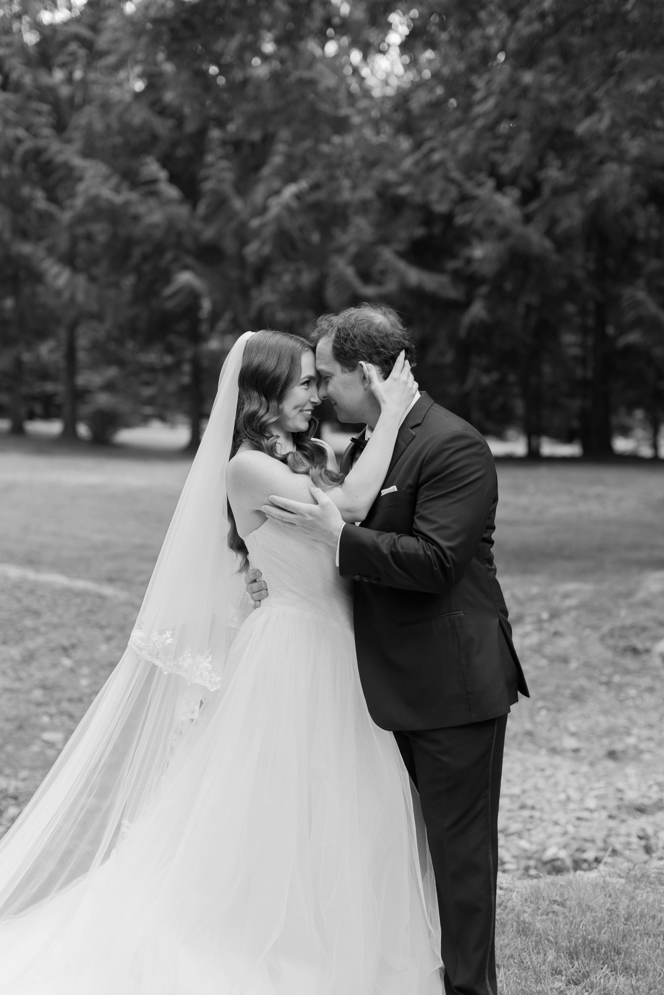 Black and white photo of a bride in a veil and gown embracing her groom in a dark suit, foreheads touching, in a park surrounded by trees.