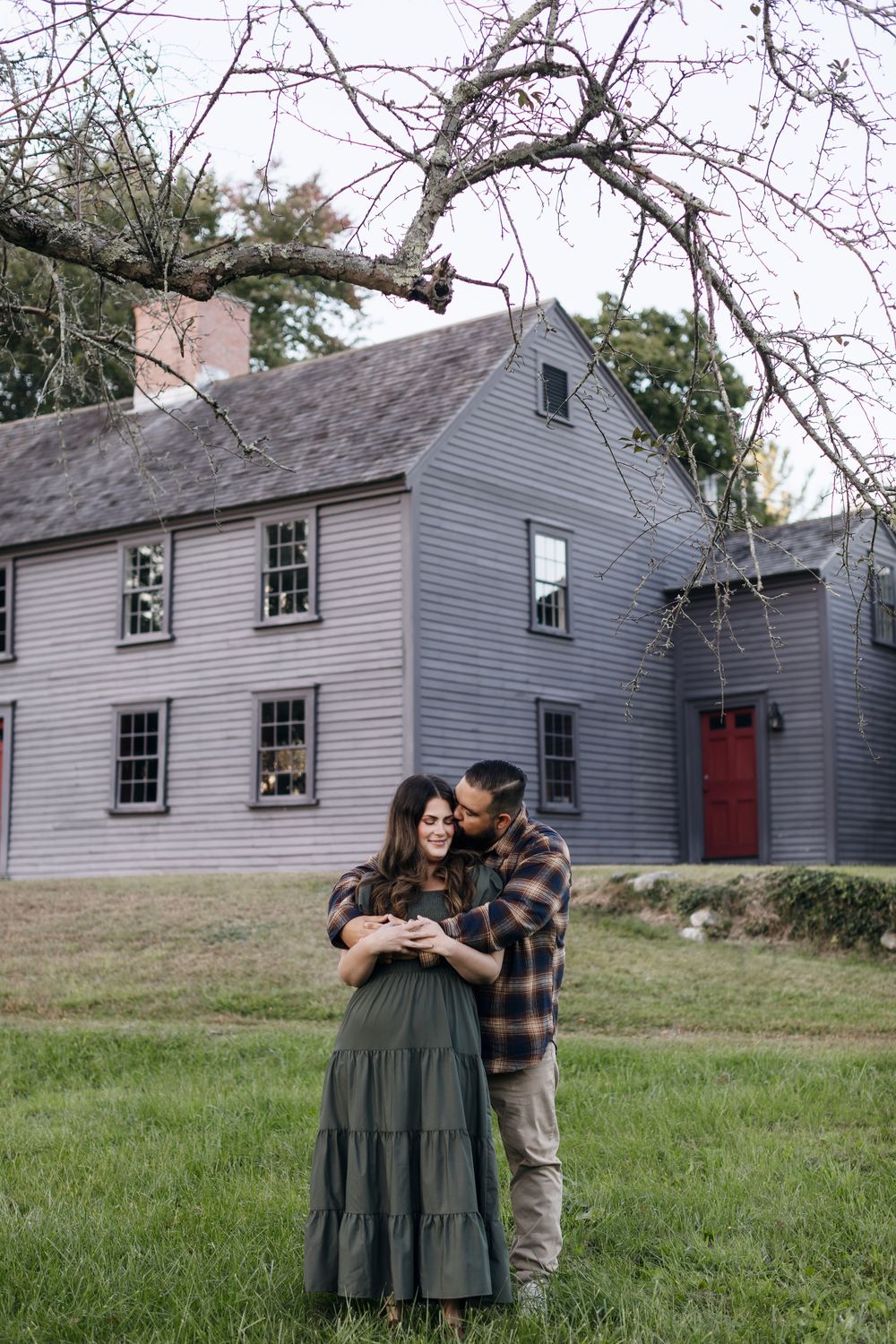 Couple embraces in front of a gray colonial house under tree branches for engagement photos in Concord, MA. engagement-photographer-Concord