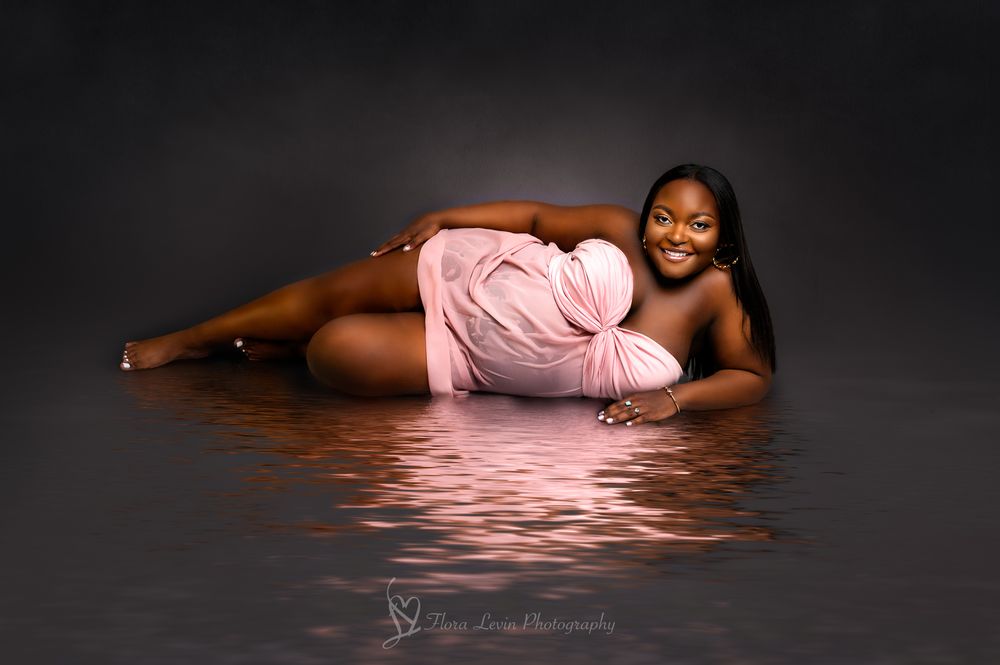 Pregnant woman  smiling wearing a pink shear wet dress, laying on her side by a shiny water with her reflection against a gray background.