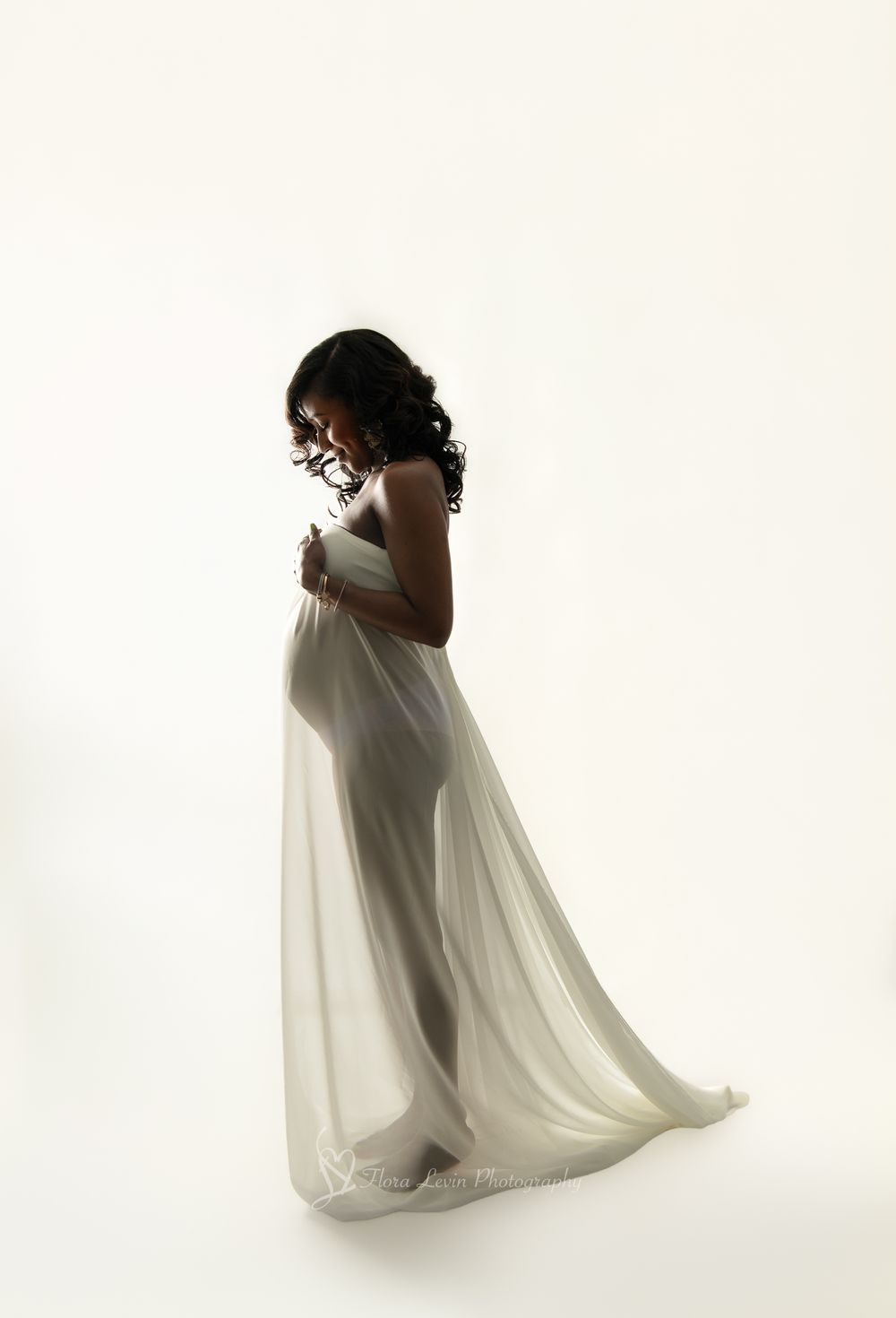Pregnant woman in flowing white chiffon gown cradling baby bump, elegant maternity photography on white background. by Flora levin.