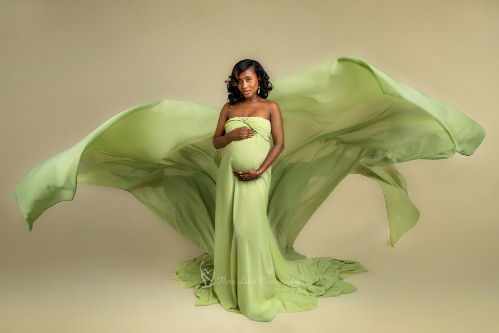 Pregnant woman in flowing green maternity gown with dramatic fabric wings, posing gracefully against neutral background.