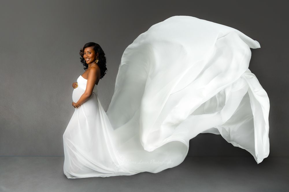 Pregnant woman in flowing white gown with fabric billowing gracefully against gray studio background.