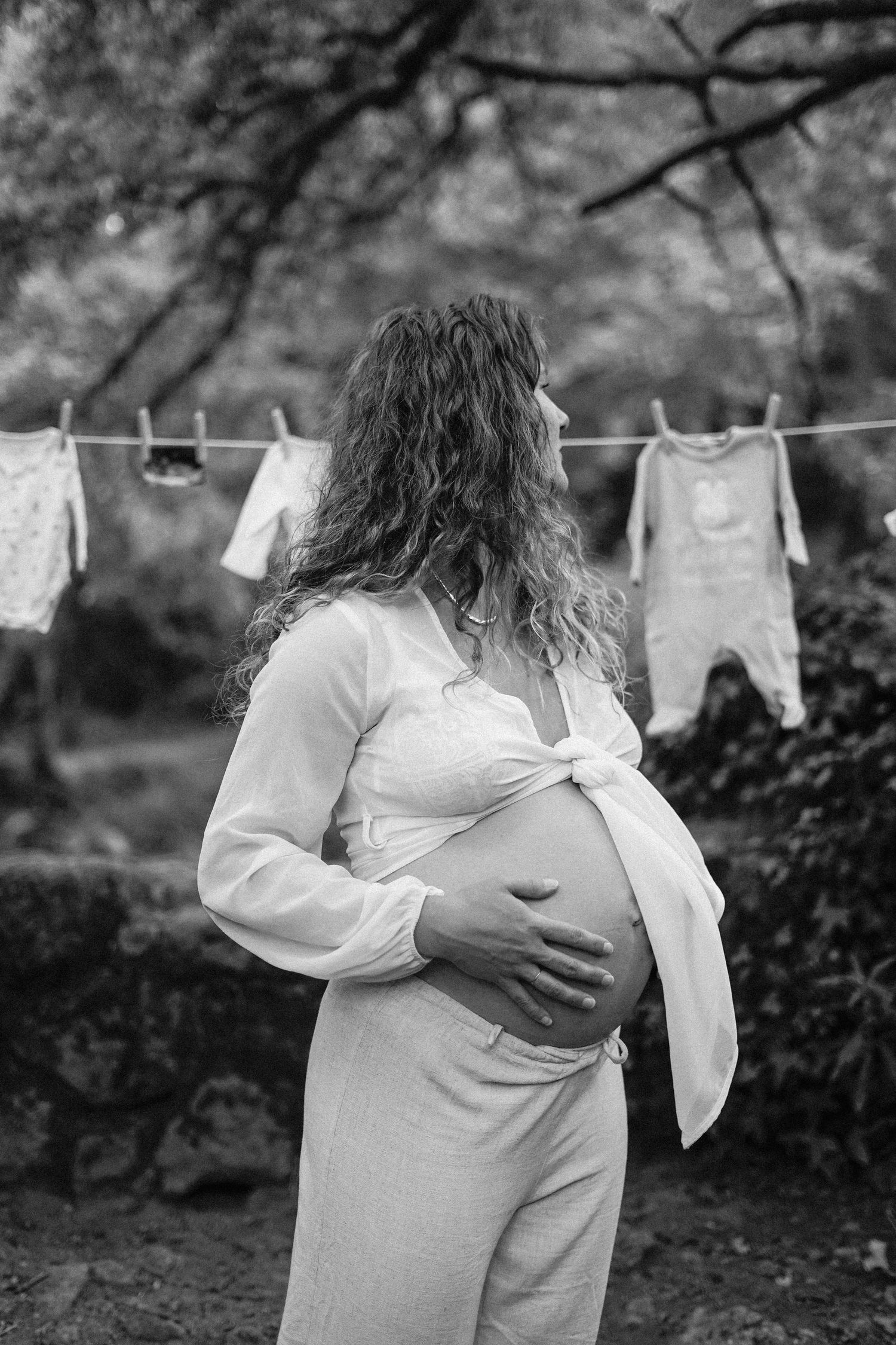 Maternity photo of expectant mother by clothesline with baby garments in garden setting.
