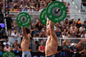 Two male CrossFit athletes perform overhead barbell lifts with Rogue weight plates at an outdoor competition.