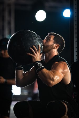 Muscular man in black tank top lifting heavy medicine ball during intense CrossFit workout in dark gym.