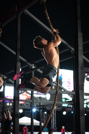Shirtless male athlete climbing a thick rope during a nighttime CrossFit competition under bright arena lights.