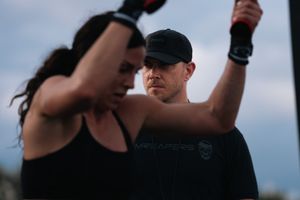 A man in a black cap coaches a woman during an outdoor fitness training session at dusk.