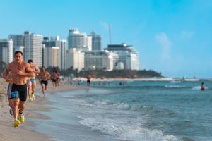 Shirtless runners jogging along a sunny Miami Beach shoreline with luxury high-rise buildings in the background.