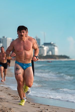 Muscular man running on a beach in swim briefs during an outdoor fitness competition.