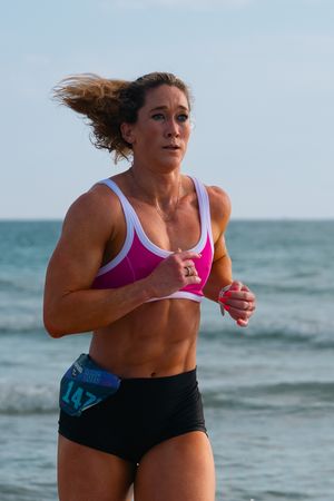 Fit woman wearing race number 147 running on beach in pink sports bra and black shorts during seaside race.