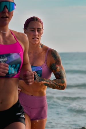 Two tattooed women in pink athletic wear run a beach race, competing in a triathlon event by the ocean.