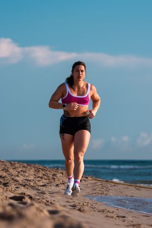 Woman in pink sports bra and black shorts running along a sandy beach on a sunny day.