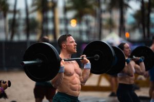 Athletic man lifting barbell outdoors on beach during CrossFit workout with palm trees in background.