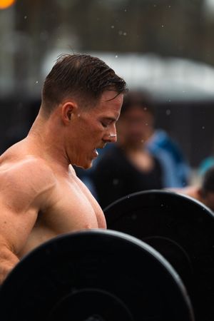 Shirtless muscular man lifting a barbell outdoors in the rain during a fitness competition.
