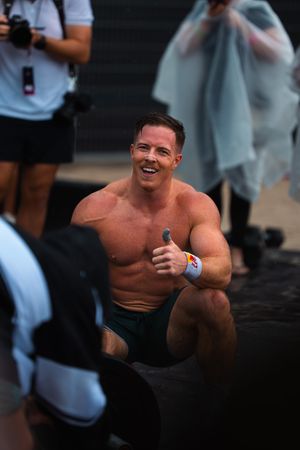 Shirtless muscular man smiling and giving thumbs up at outdoor fitness competition.