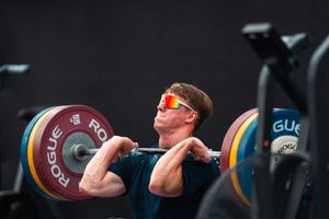 Male athlete performing a clean and jerk lift with Rogue barbells wearing orange sunglasses.