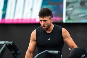 Muscular male athlete in black Adidas sleeveless shirt training on exercise bike at fitness event.