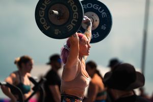Female CrossFit athlete with colorful hair lifting Rogue barbell overhead at outdoor competition.