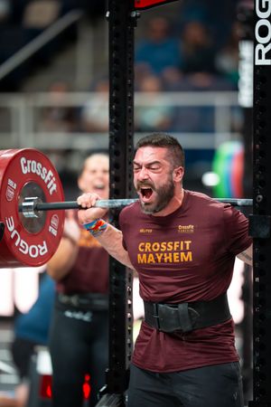 CrossFit athlete performing a powerful barbell snatch movement in a maroon competition shirt.
