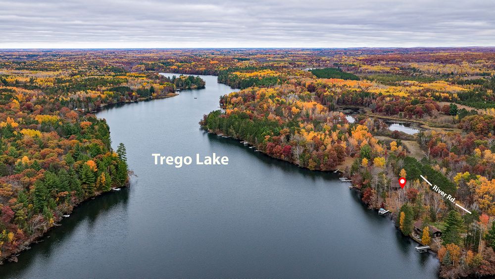 Aerial view of Trego Lake surrounded by forest in vibrant orange, red and yellow autumn foliage under a soft cloudy sky.